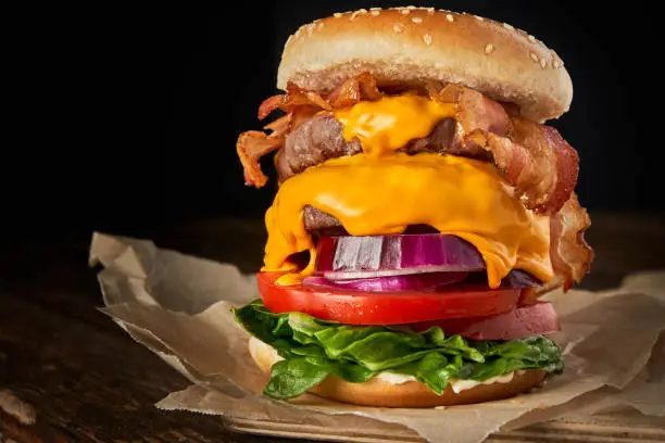 Close-up of home made tasty burger with onion, lettuce, cheddar cheese, bacon and tomato on a wooden table, with copy space.Food concept