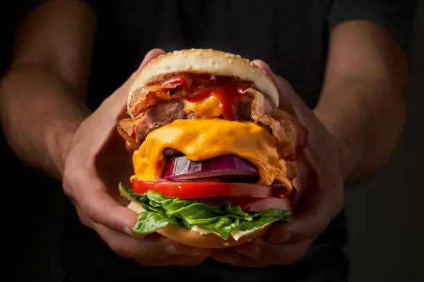 Photo of Unrecognizable young man holding in hands fast burger on black background