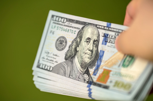 Close-up of a hand holding a bundle of US $100 bills. Green background. Shallow depth of field.