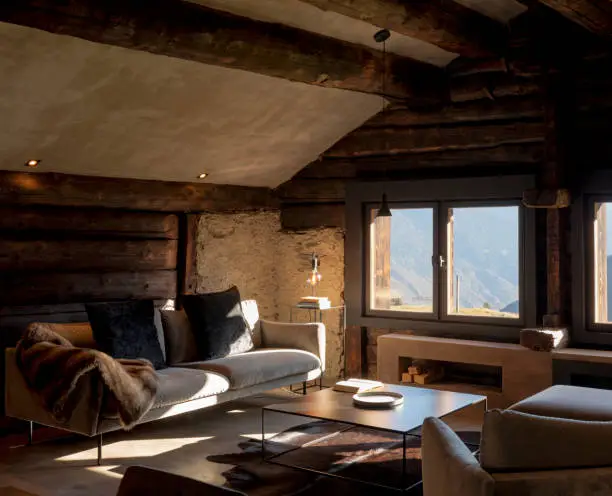 Mountain chalet living room with large sofa, two armchairs and a large window overlooking the Alps. Wooden beams and nobody inside.