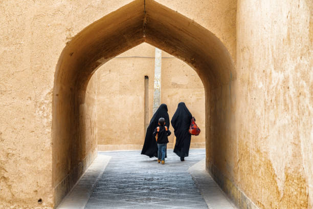 Iranian women wearing black chador walking along street of Yazd Yazd, Iran - 27 October, 2018: Iranian women wearing black chador walking along narrow street of the historical city of Yazd. Unique Persian architecture of the ancient town. islamic architecture stock pictures, royalty-free photos & images