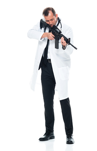 Front view of aged 40-44 years old with brown hair caucasian young male doctor standing in front of white background wearing lab coat who is angry who is shooting a weapon and holding weapon and using gun
