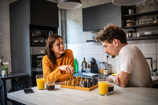 A young beautiful couple is playing chess in their kitchen while drinking their morning coffee, using their free day to relax.