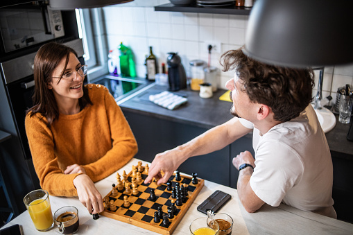 A young beautiful couple is playing chess in their kitchen while drinking their morning coffee, using their free day to relax.