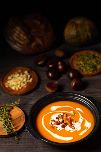 Pumpkin and chestnut soup with cream, pork belly and thyme aside seeds and thyme, chestnuts and bread on a vintage dark wood background. Vertical view. stock photo