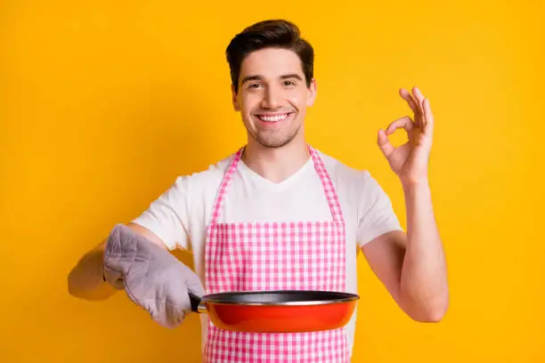 Photo portrait of guy in kitchen gloves showing ok-sign holding frying pan isolated on vivid yellow colored background.