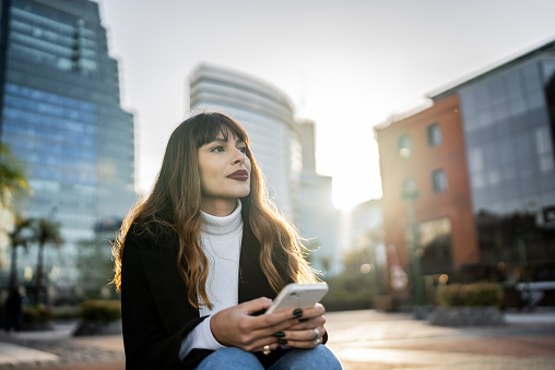 Young woman sitting outdoors holding smartphone and contemplating