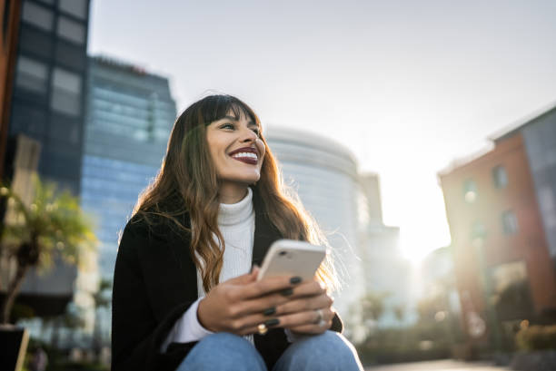 屋外に座ってスマートフォンを持ち、熟考している若い女性 - satisfaction clothing argentina smiling ストックフォトと画像