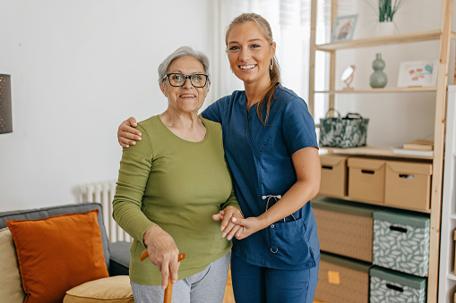 Female caregiver helping and supporting senior patient to walk at home, senior women is standing and holding walking cane