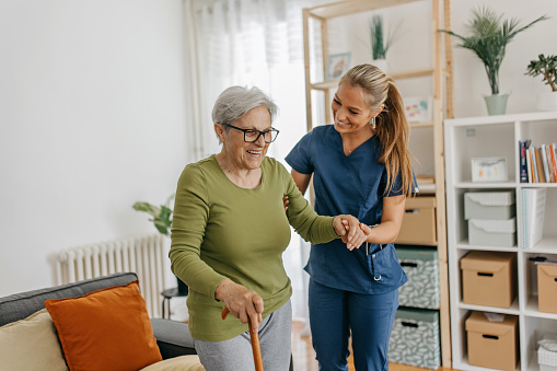 Female caregiver helping and supporting senior patient to walk at home, senior women is standing and holding walking cane