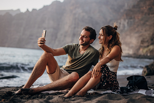 Young couple sitting on the beach at sunset and taking a selfie