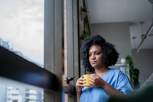 Black nurse looking out the window with a cup of coffee in her hand