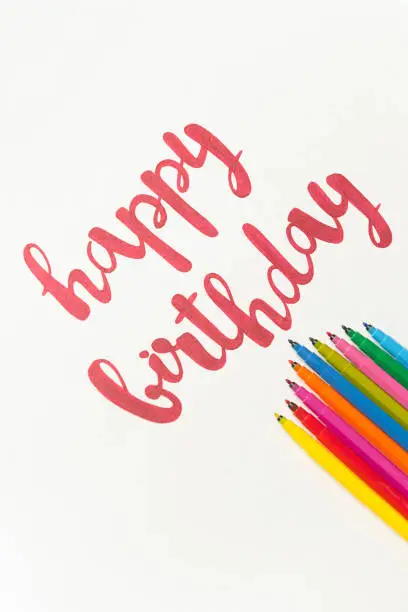 Inspirational phrase 'Happy birthday' for greeting cards and posters drawing with red marker on white paper. Top view of lettering, bunch of colourful markers