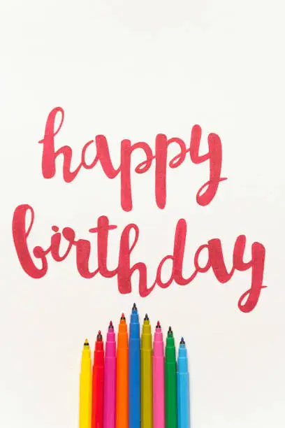 Colourful phrase 'Happy birthday' for greeting cards and posters drawing with red marker on white paper. Top view of lettering, bunch of colourful markers