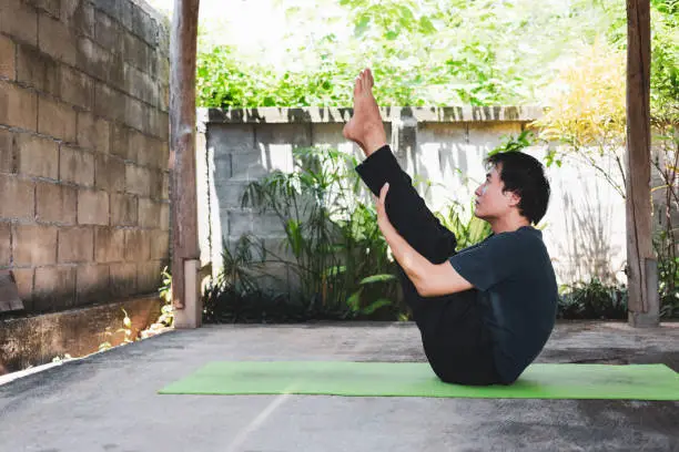 Photo of Healthy living concept of young Asian man practicing yoga asana Paripurna Navasana Pose or Full Boat Pose, work out and poses on a green yoga mat. outside exercise in the garden. healthy lifestyle.