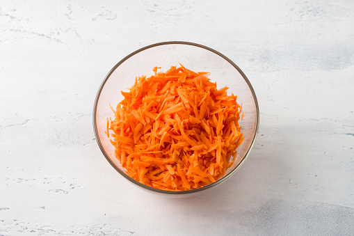 Glass bowl with grated carrots on a light gray table, top view. Cooking homemade healthy food