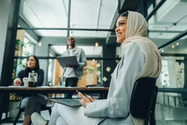 Happy Muslim businesswoman holding wireless technology while sitting in an office meeting. Cheerful young businesswoman wearing a hijab in an inclusive workplace.