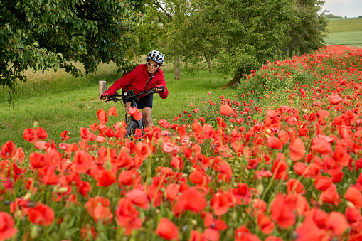 nice, active senior woman having fun on her electric bicycle in a huge field of blooming red poppies