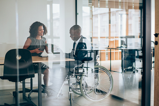 Shot of an african-american businessman in wheelchair having discussion with his female colleague in the board room. Photo of elegant businesswoman working with her disabled colleague in the office. Corporate business persons discussing new project and sharing ideas in the workplace. The view is through glass.