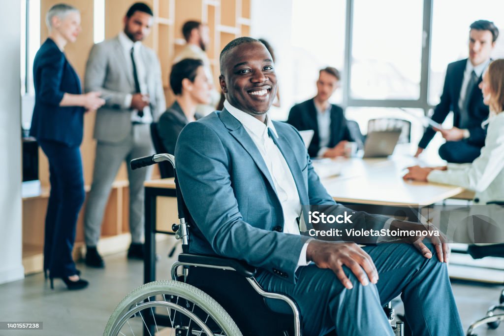 Portrait of smiling disabled businessman in wheelchair at meeting. Shot of an african-american businessman in wheelchair at business meeting in the board room. Paralyzed man in a wheelchair at meeting with his colleagues in the office. Equality Stock Photo