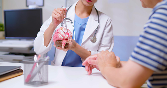 closeup of attractive young brunette ponytail female asian doctor wear white coat and stethoscope pen pointing explaining brain model to gray hair old sick man at clinic