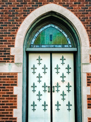 Front entrance to a United Methodist church with ornate doors. Fleur de lis symbols adorn the front doors. Arched doorway with stained glass.