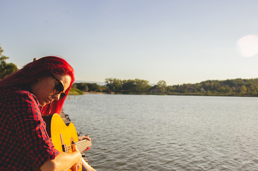 Blonde woman  playing guitar besides a lake