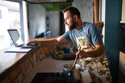 Bearded Male Working On Laptop And Making Dinner At Same Time