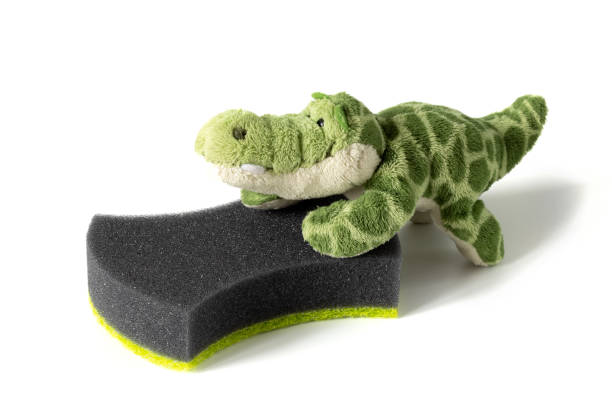 istock Toy plush crocodile pushes black sponge for household needs on white background. Concept of involving children in housework, surprise for mom, cleaning kindergarten, animal care 1401576839