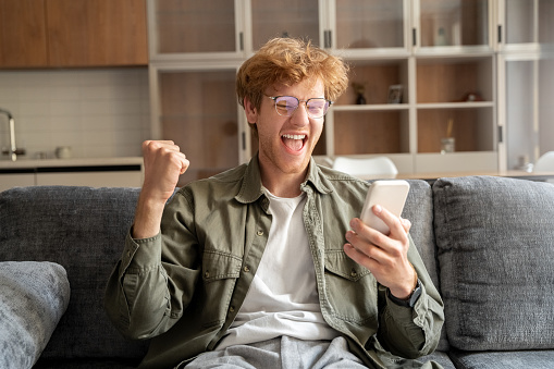 Happy overjoyed excited young irish ginger man in glasses gesturing and screaming yes to mobile webcam. Unexpected reward or success, lottery win, shock news, giveaway or online bet gambling victory