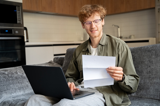 Young ginger irish freelance man working with laptop and document at home office. Millennial employer reading paper CV doing remote recruitment work. Redhead guy paying bill tax online