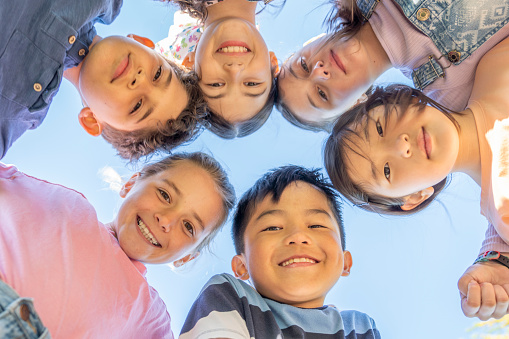 A small group of ethnically diverse school aged children huddle together in a circle with their heads pressed together as they pose for a portrait.  They are each dressed casually and are smiling in this ground view photo.