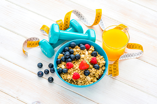 Healthy lifestyle concept: granola and berries bowl, orange juice, dumbbells and tape measure shot from above on white table. High resolution 42Mp studio digital capture taken with Sony A7rII and Sony FE 90mm f2.8 macro G OSS lens