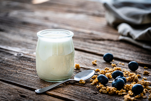 Close up view of a glass container filled with fresh healthy yogurt shot on rustic wooden table. A spoon and berry fruits are around the container.