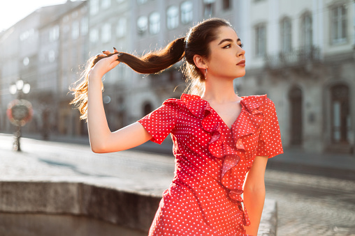Brunette girl in red dress holding her long hair tail. Young woman summer portrait in morning light