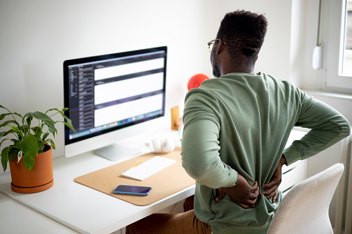 Male freelancer having back problems while sitting at his desk in front of the monitor, expressing his pain by holding his back with his hands