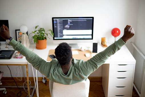 Rear view of a male freelancer enjoying the comfort of his home office, sitting at his work desk, celebrating a job well done in front of his desktop PC by holding his hands up in the air