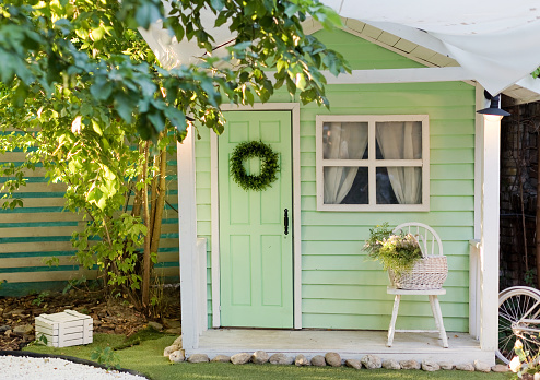 Small green shed garden house with window, door and chair outside. Kids playhouse, garden summerhouse background