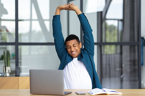Young African American businessman man working, sitting at the workplace in the office stretching his back at desk