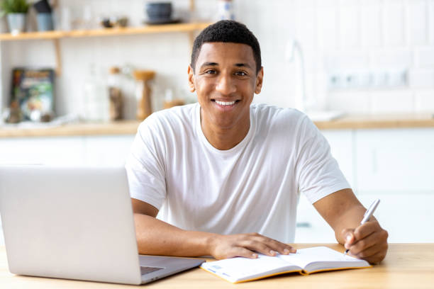 Smiling African american male student or freelancer studying remotely from home, using a laptop looks at the camera and smiles friendly. Online lesson, e-learning at home concept stock photo