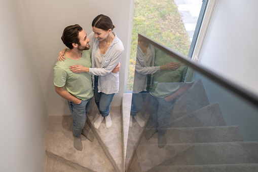 Happiness. Top view of happy young adult man and woman standing hugging looking at each other on stairs at home