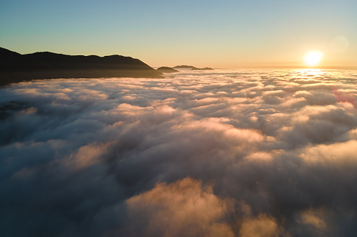 Aerial view of colorful sunrise over white dense fog with distant dark silhouettes of mountain hills on horizon.