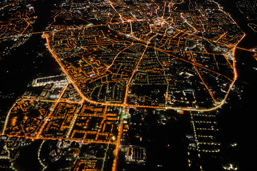 Aerial view from airplane window of buildings and bright illuminated streets in city residential area at night. Dark urban landscape at high altitude.