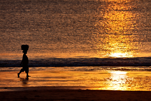 African woman walking home from work in sunset on beach carrying goods on her head.
