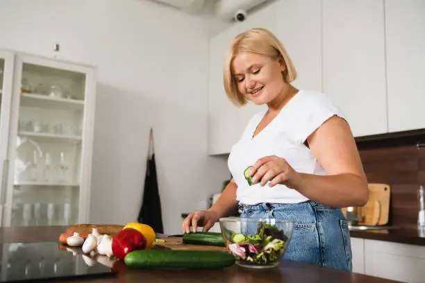 Photo of Young caucasian plump plus size woman cooking making salad, healthy food, dieting, counting calories, preparing dinner lunch at home kitchen