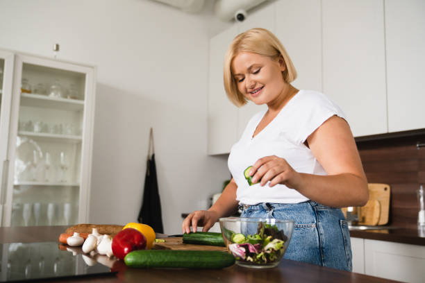 jovem mulher caucasiana gorda plus size cozinhando salada, comida saudável, dieta, contando calorias, preparando o almoço do jantar na cozinha de casa - large build - fotografias e filmes do acervo