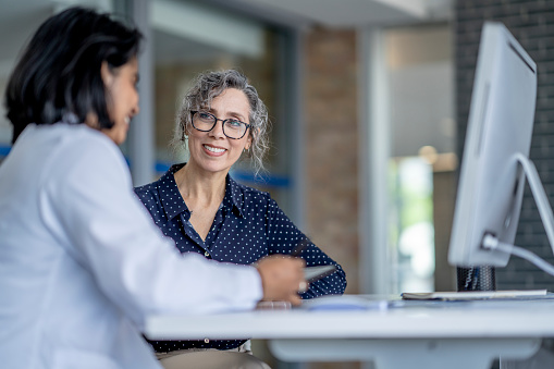 A middle aged woman sits with her female doctor of Middle Eastern decent as they review her medical concerns.  She is dressed semi-casually and sharing with the doctor her recent discomfort.  The doctor is wearing a white lab coat and taking notes on her tablet as the two talk.