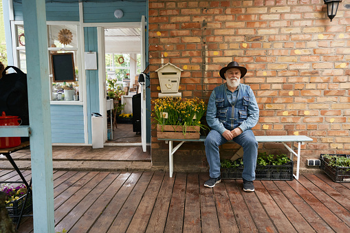Full length portrait of aged male sitting on bench with boxes plants for sale at entrance to rural boutique