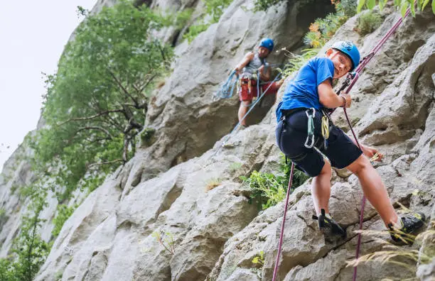 Smiling teen boy in protective helmet abseiling from cliff rock wall using rope, Belay device and climbing harness with father in Paklenica Park Croatia. Active extreme sports time spending concept