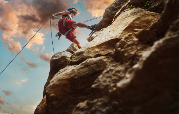 istock Muscular climber man in protective helmet abseiling from cliff rock wall using rope Belay device and climbing harness on evening sunset sky background. Active extreme sports time spending concept. 1401610146